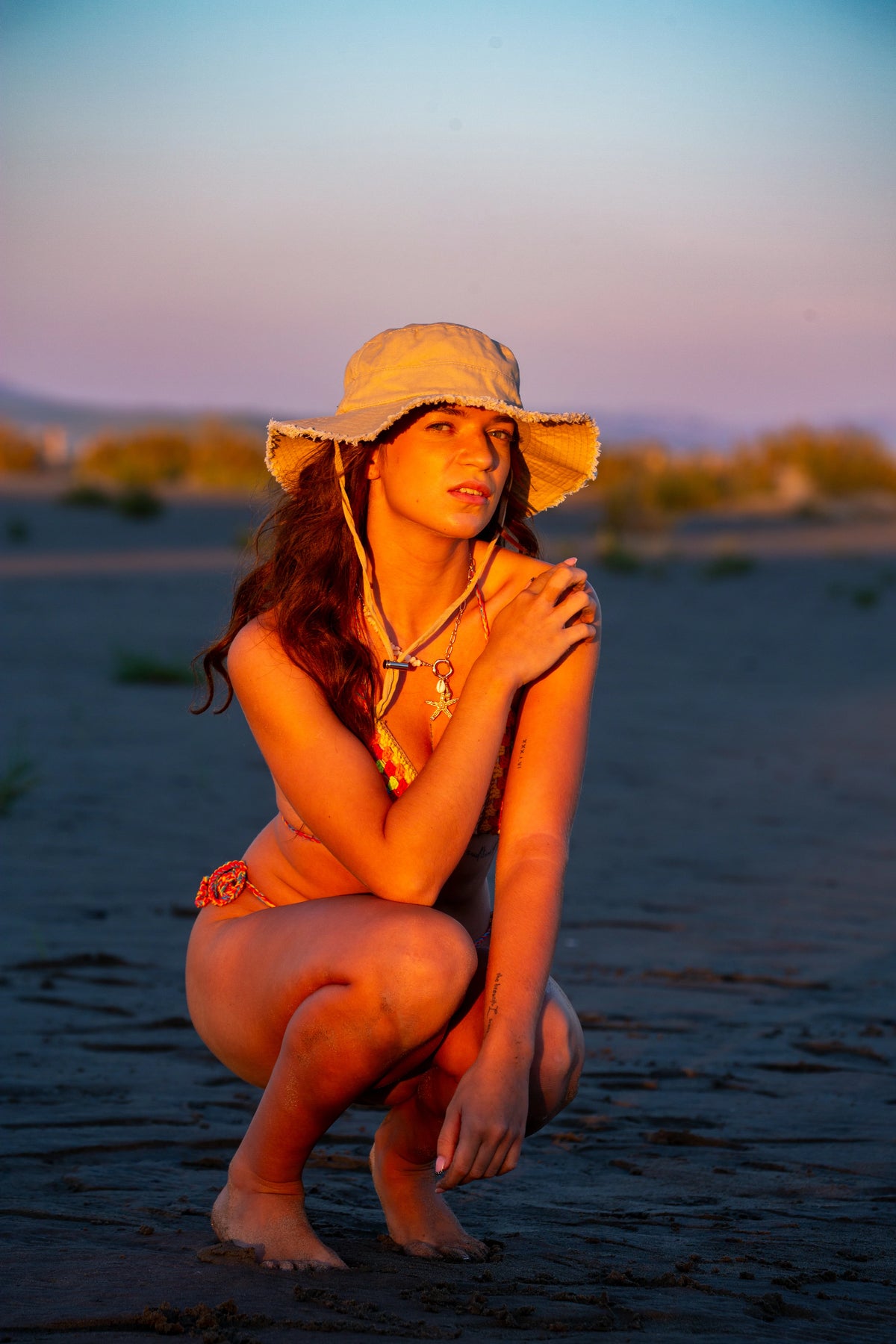 Jeune femme debout sur la plage, ajustant son chapeau de paille, portant le maillot de bain "Sienna" en crochet multicolore façon patchwork (granny square) Soleil Leblon